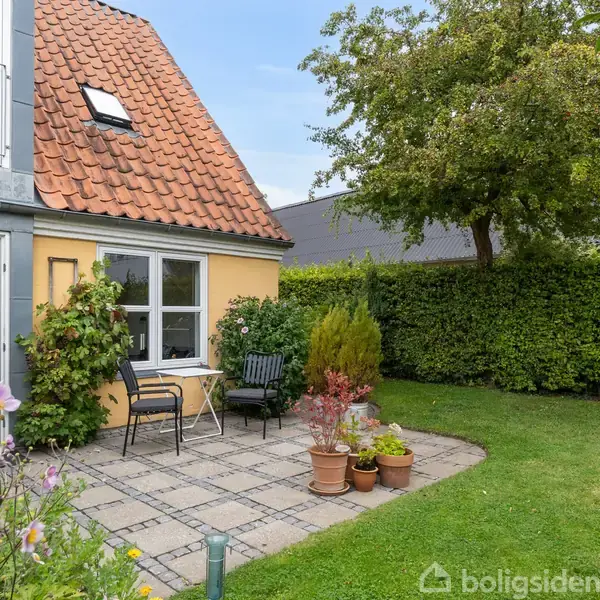 Et gult hus med rødt tegltag, et vindue og en terrasse med borde og stole i en have. Der er græsplæne, træer og blomster omkring.