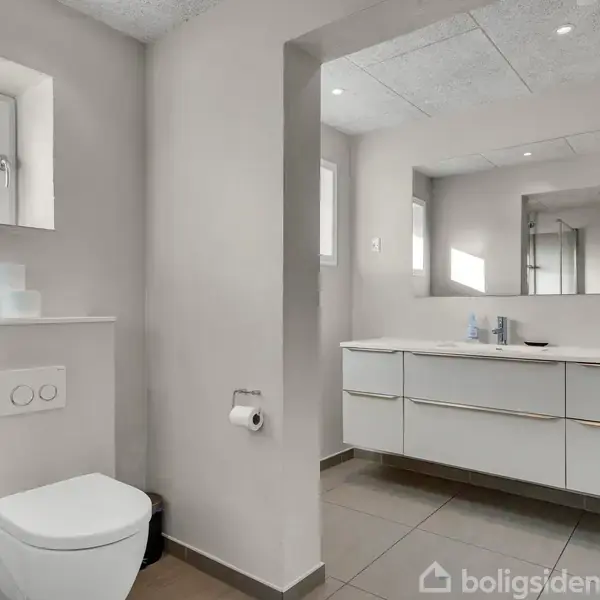 Toilet og skabsarrangement i moderne badeværelse med spejl. Handlingsfri kontekst, lyse fliser og vægge med spotlys i loftet. Rullegardin i vindue ved siden af toilettet.