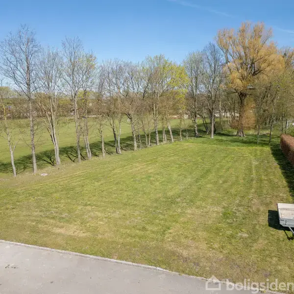 Græsplæne strækker sig over et åbent område, omkranset af træer og en hæk. En lille trailer står stationær til højre. Himlen er klar og blå.