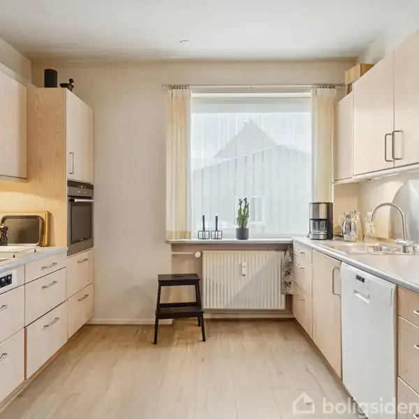 Et lyst køkken med træskabe, hvor en elkedel står på komfuret. Der er en ovn indbygget i højskabet. Et vindue med gardiner og planter udsmykker baggrunden. En stol står ved radiatoren.