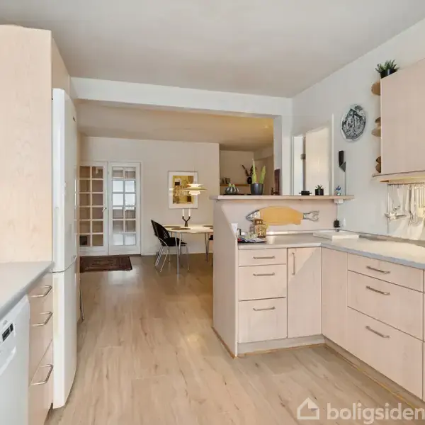 Køkken med lyse træskabe og indbygget opvaskemaskine; køkkenredskaber hænger under overskabene. I baggrunden ses en spisestue med en lampe over bordet og dekorationer på væggene.