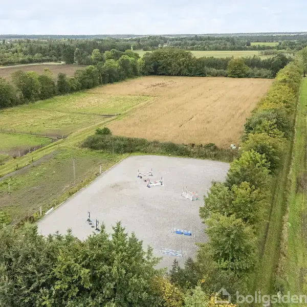 En rektangulær grusbane er omgivet af grønne marker og træer. Enkelte mennesker er spredt på banen, der tilsyneladende bruges til en udendørs aktivitet.