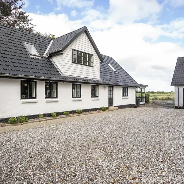 Et hvidt landhus med sort tegltag står ved en grusbelagt gårdsplads. Et separat garage- eller udhus ligger til højre. Træer og marker ses i baggrunden.