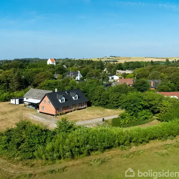 Hus med mørkt tag omgivet af frodig natur og græsplane, beliggende i en landlig landskabsceneri med grønne marker og spredte træer, set fra en luftig vinkel.