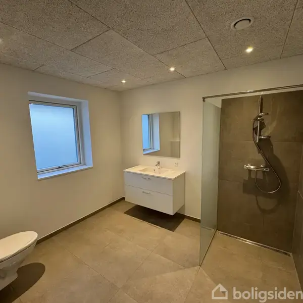 Badeværelse med toilet, håndvask og bruser. Håndvasken er under spejlet. Rummet er lyst med flisegulv og et vindue. Brusenichen har glasvægge.