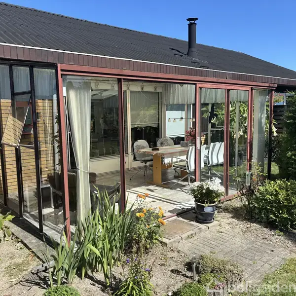 Et glashus med røde rammer står i en have. Indenfor ses et bord og stole. Omgivet af grønne planter og blå himmel.