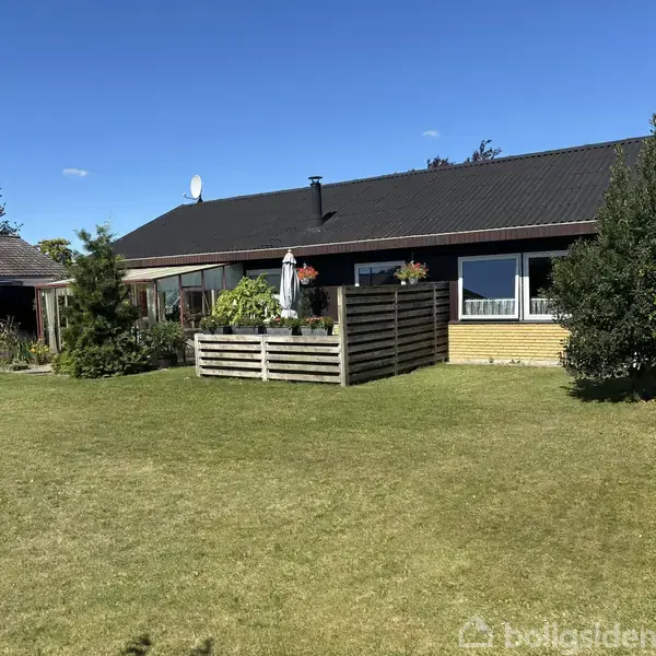 En etplanshus med sort tag står i en have med græsplæne, træer og en terrasse med blomsterkrukker. Solen skinner, og en garage med bil ses til venstre.