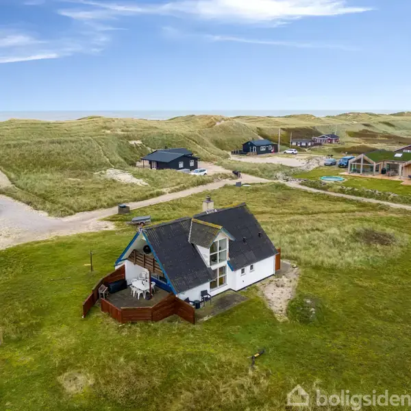 Et bindingsværkshus med stråtag står alene på en grøn mark omgivet af klitter. Flere huse kan ses i baggrunden, placeret blandt sandklitterne under en blå himmel.