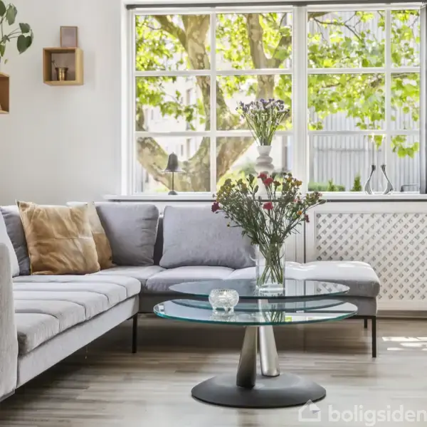 Sofa står i stuen; grå L-form, pyntet med puder. Rundt glasbord foran med blomster i vase. Store vinduer bagved med udsigt til grønne træer.