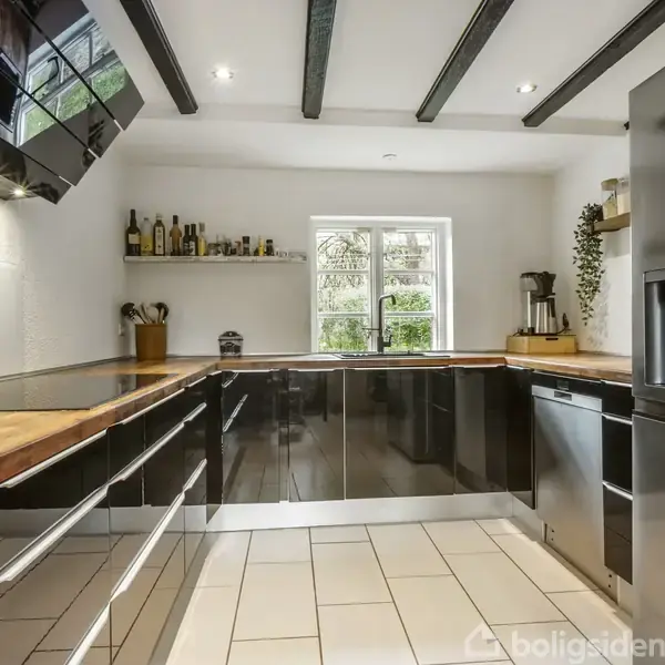 Et moderne køkken med sorte skabe og træbordplader, stiliseret med indbyggede loftlamper. Køkkenet indeholder en stor grå køleskab og et vindue, der åbner mod lyse omgivelser. Skabe er fyldt med flasker og krydderier.