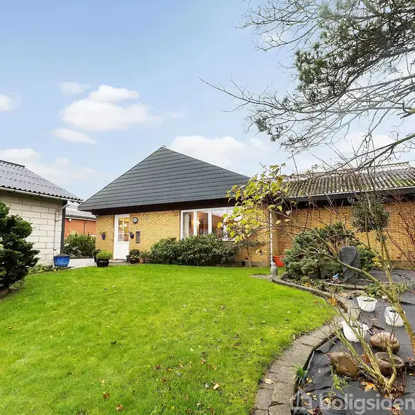 En gul murstensvilla står i en velplejet have med grøn græsplæne omkranset af buske og træer. Himmel er blå, og der er en flisebelagt sti til højre.