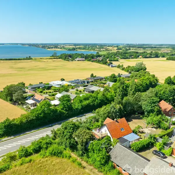 Huse med røde tage står stille omgivet af frodige grønne træer og gyldne marker; baggrunden viser en stille sø under en klar blå himmel.