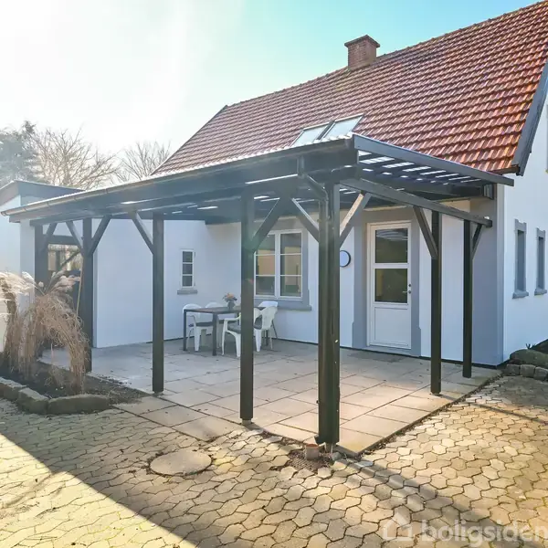 Et hvidt hus med rødt tegltag står på en flisebelagt terrasse, hvor to stole og et bord er placeret under en pergola. Omgivet af en belagt indkørsel og grønne buske.