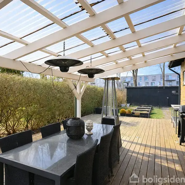 En udendørs terrasse med et tag af gennemsigtig plast. Et spisebord med sorte stole står under taget. En grill og terrassevarmer er til højre, omkranset af hække.