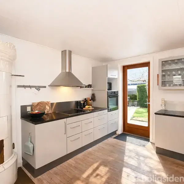 Et moderne køkken med hvide skabe og sorte bordplader, en emhætte over kogepladen. En rund hvid kakkelovn til venstre. En dør fører ud til en haveområde.