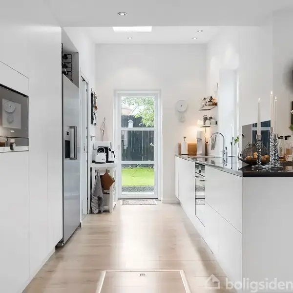 Et moderne køkken med hvide skabe og apparater. Køleskabet står til venstre. Vindue og dør i baggrunden fører ud til en have med grønt græs. En stor sort ur hænger på væggen. Blomster og dekorationer står på køkkenbordet til højre.