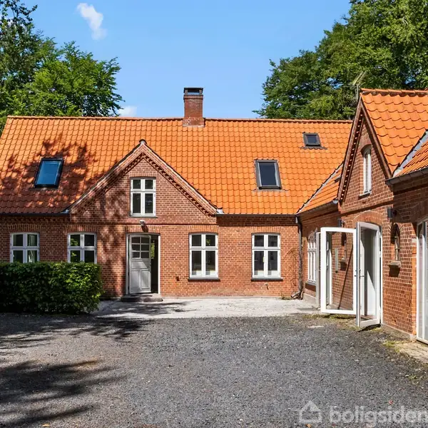 Et rødt murstenshus med orange tegltag står i en skovklædt omgivelser. Døre og vinduer er hvide, og gårdspladsen er dækket af grus.