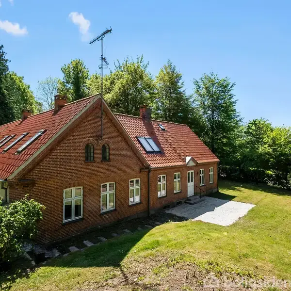 Rødt murstenshus med skråtag, mange vinduer og antenne; omgivet af græsplæne og træer under en blå himmel.
