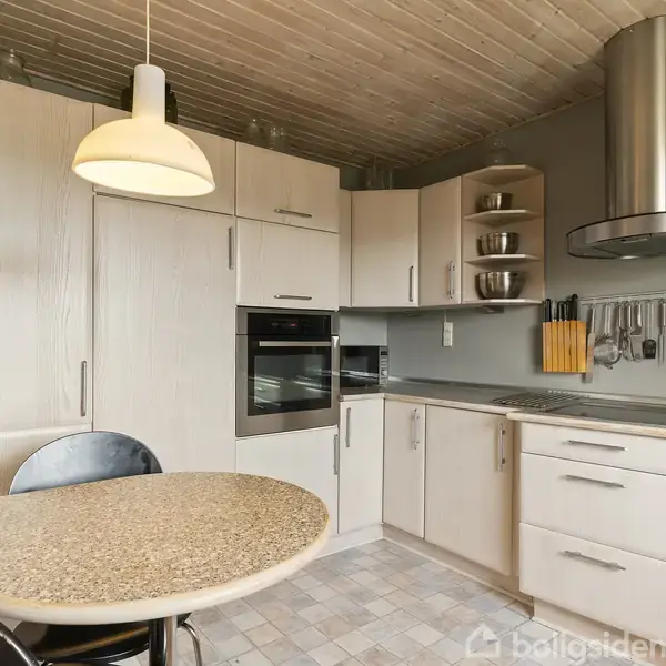 Et lyst køkken med et rundt spisebord, ovn indbygget i skabsvæg, samt hylde med metalskåle. Køkkenredskaber hænger over en komfurhjørne. En stor emhætte er ovenover komfuret.