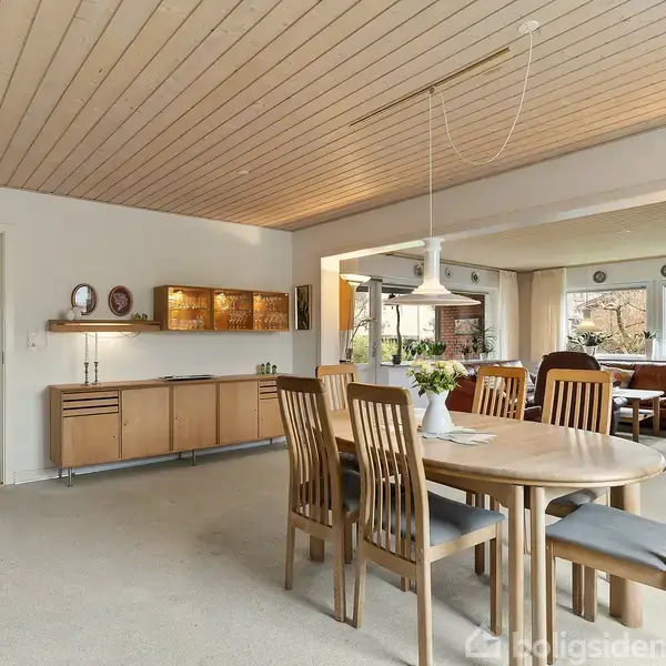 Rundt spisebord med seks stole står i et lyst, rummeligt stueområde. Vinduer giver udsigt til haven udenfor. En skænk og væglamper supplerer indretningen, mens lofterne er i træ.