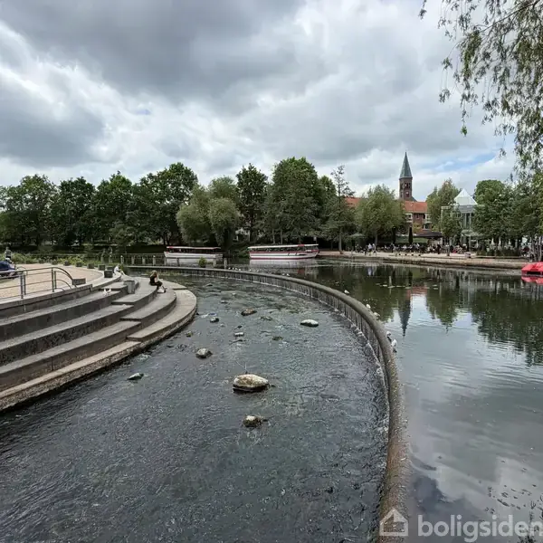 Trapper fører ned til vandet i en rolig bypark med en flod, der flyder forbi. Træer og en kirke ses i baggrunden under en overskyet himmel.