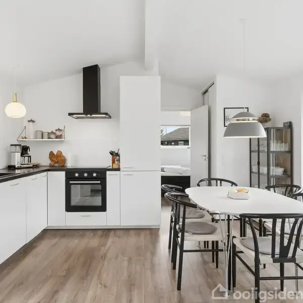 Køkken-alrum med hvidt køkken, sorte stole og et spisebord. Der er moderne apparater, trælaminatgulv, store vinduer og hvidmalede vægge. Rummet har to hængelamper og en dør til et tilstødende rum.