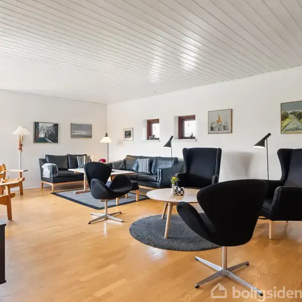 Stue med sorte lænestole og sofaer omkring et rundt bord, omgivet af lyse trægulve og hvide vægge med kunst. Vinduerne giver udsigt til grønt udenfor.