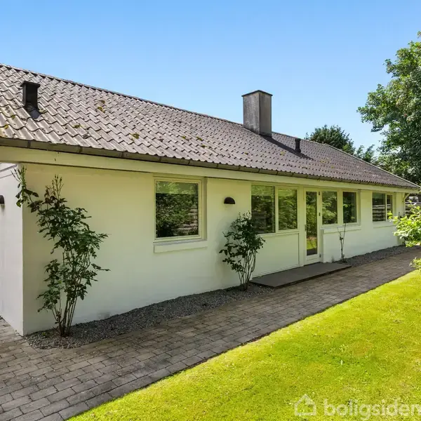 En hvid, enetages villa med gråt tegltag står stille i en solrig have omgivet af grønne planter og en velplejet græsplæne. Fliser danner en gangsti langs huset.