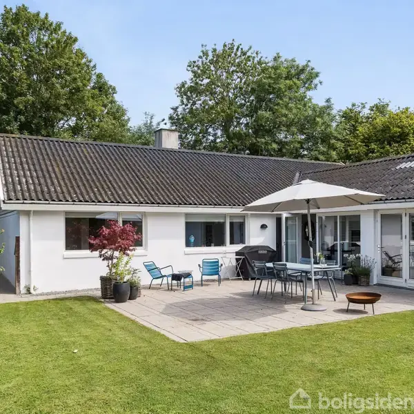 Hus med hvid facade, har terrasse med parasol og møbler på stenbelægning. Græsplæne omgiver terrassen, mens træer i baggrunden skaber en naturlig ramme.