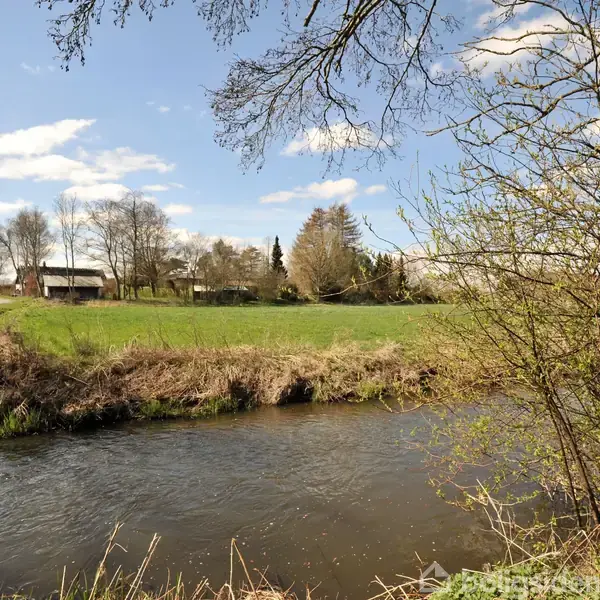 En flod strømmer stille gennem landskabet, omgivet af græs og buske. I baggrunden er marker og et lille hus synligt under en blå himmel med spredte skyer.