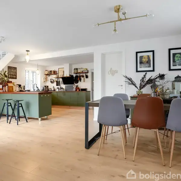 Spisebord med fire stole står i et lyst, åbent køkken-alrum. Et moderne køkken med grønne skabe og barstole ses i baggrunden. Trappe til venstre og kunstværker på væggen.