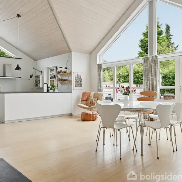 Lyst åbent køkken-alrum med højt til loftet, spisebord med stole og store vinduer mod terrasse og have. Køkkenet er moderne med grå og hvide nuancer. Ingen tekst synlig.