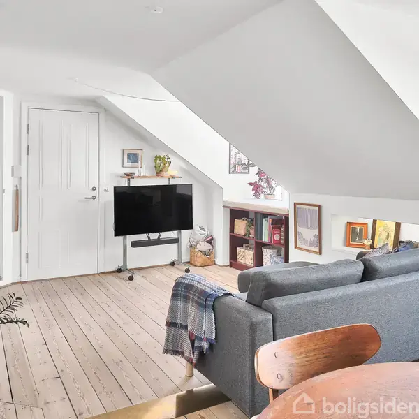En grå sofa står i en lys loftstue med skrå vægge, placeret foran et lille, sort TV. Omgivet af træmøbler, planter og en vaskesøjle i et moderne, rummeligt miljø.