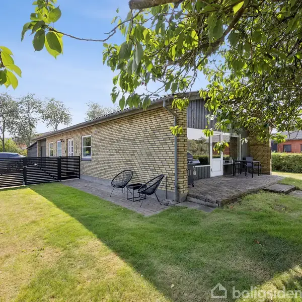Et enkelt, ét-etagers hus med gul murstensfacade, der står på en velholdt græsplæne. Foran huset er en terrasse med stole og et bord. Omkring er der træer og hække.