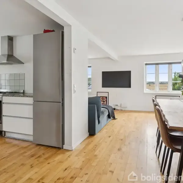 Køkken-alrum med trægulv. Til venstre er et moderne køkken med stålkomfur og emhætte. Til højre står spisebord med stole og en vase med blomster. Stue i baggrunden.