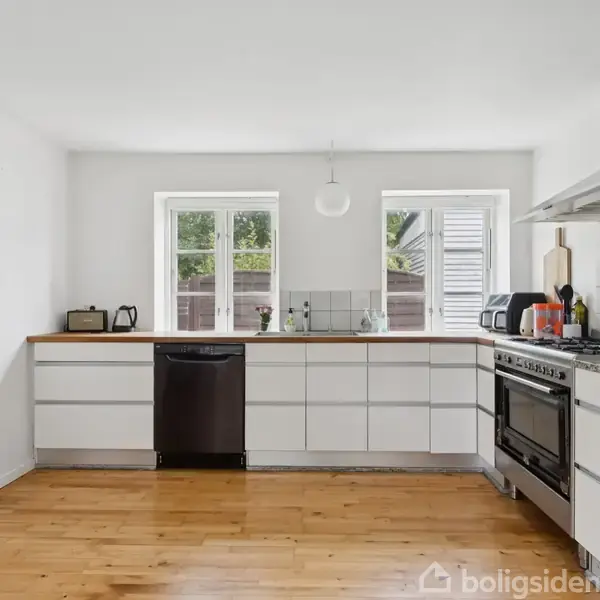 Et moderne køkken med lyse skabe og træbordplade. En opvaskemaskine og ovn står mellem skabene. To vinduer lukker naturligt lys ind. Treetage og hvide vægge omgiver rummet.