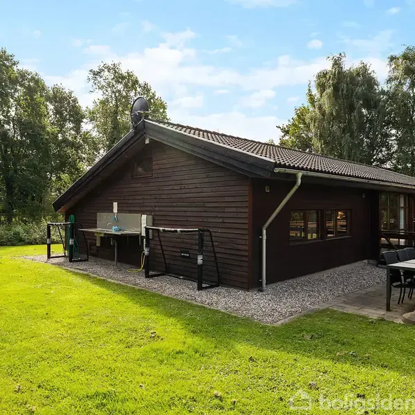 Et træhus med mørkebrune vægge står stille i en grøn have. Et bord med stole er placeret på terrassen ved siden af. Omgivet af træer.