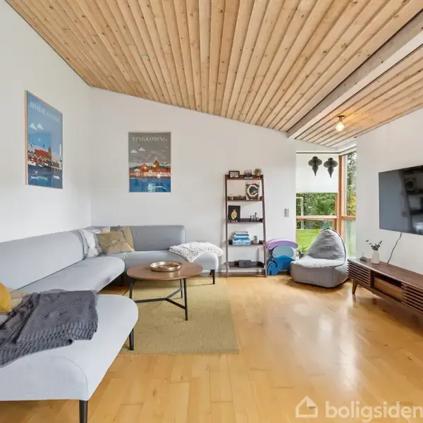 En stue med en L-formet grå sofa fyldt med puder og tæpper. Foran står et rundt sofabord. Væggene er hvide med plakater, og et stort vindue slipper lys ind.