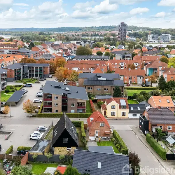 Bygninger står stille i et boligområde med røde tage og grønne haver, omgivet af veje og parkeringspladser; en rækkehuskvarter spreder sig i en bakke i baggrunden.