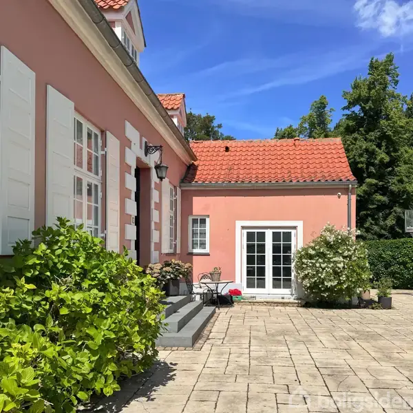 Hus med rødt tegltag og pink facade. Beliggende i en frodig have i et villakvarter omgivet af andre huse og træer.