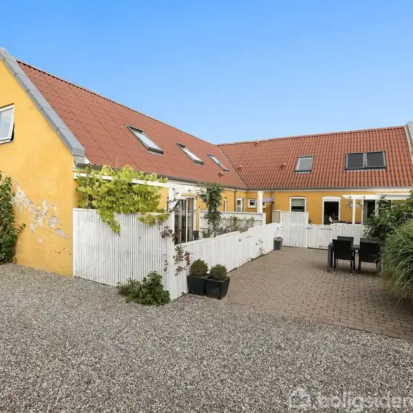 Gult hus med sadeltag og hvide vinduer. Foran huset er der en flisebelagt gårdsplads med havemøbler omgivet af planter og et lille hvidt stakit.