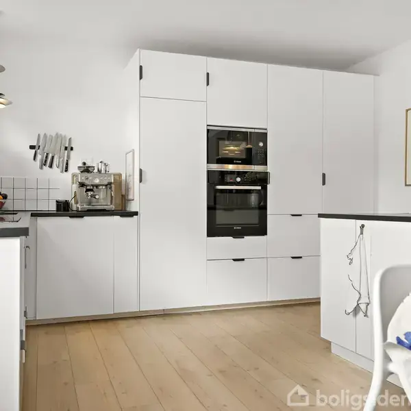 Hvidt, moderne køkken med indbyggede skabe og ovne, kaffemaskine på bordpladen. Et spisebord med blomstret dug står i forgrunden, omgivet af lysarmaturer og en vase med blomster i venstre hjørne.