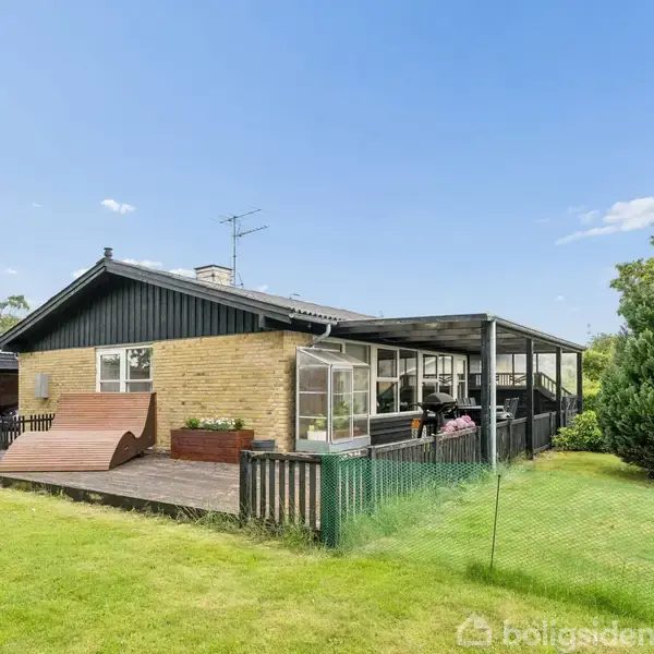 Et enkelt hus med fladt tag, brun facade og overdækket terrasse. Det ligger i en grøn have med træer og græsplæne.