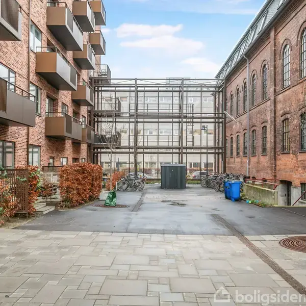 En brostensbelagt gårdhave omgivet af røde murstensbygninger. Venstre bygning har balkoner, og der er cykler samt affaldscontainere i midten. Skyfri himmel over gården.