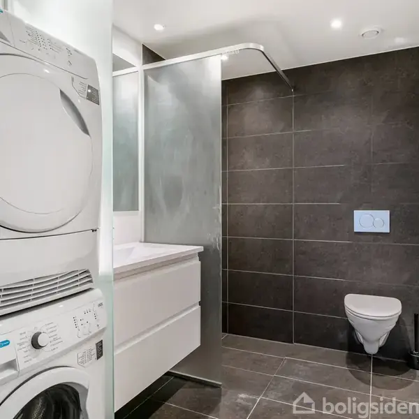 En vaskemaskine og tørretumbler er stablet i et moderne badeværelse. Ved siden af er en vask og spejl, mens brusenichen og toilet er på den modsatte væg.