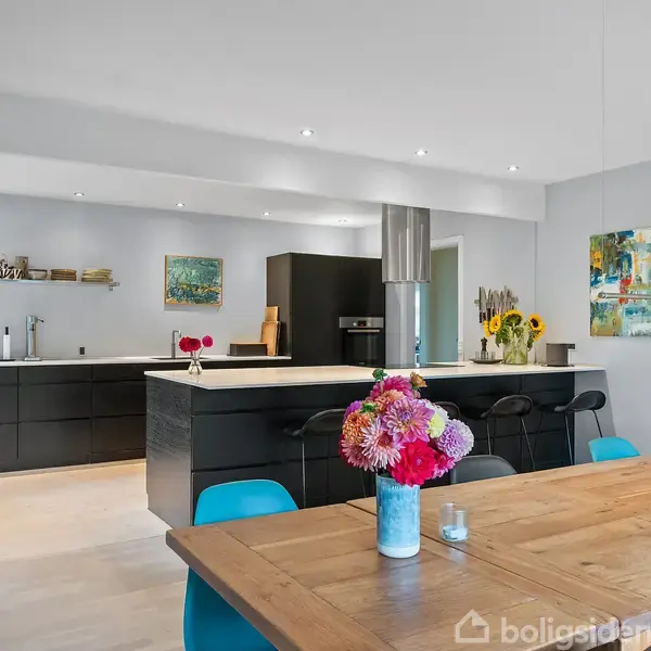 Køkken med sort, moderne inventar; blomsterdekorationer står på bordet; træspisebord med blå stole i lys og kunstnerisk udsmykket spisestue.