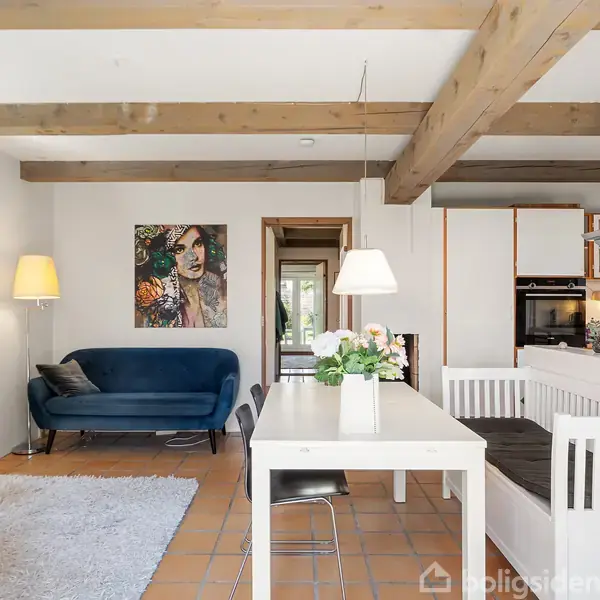 Blå sofa står langs væggen med en lampe og et maleri over. Hvidt spisebord med blomsterbuket i køkken-alrum. Træbjælker i loftet skaber hyggelig atmosfære.