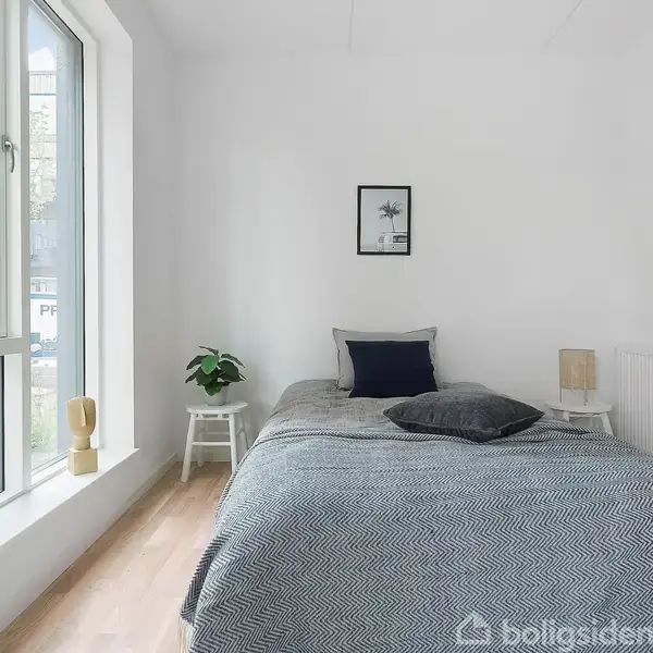 En enkeltseng dækket af et gråt tæppe står i et lyst soveværelse med hvide vægge. Ved siden af sengen ses to små natborde med planter og en lampe. Et stort vindue giver udsigt til en bymæssig baggrund.