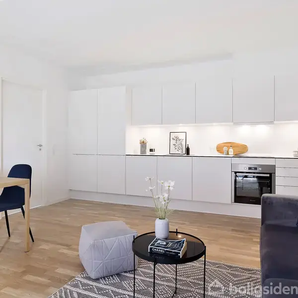 Køkken-alrum med hvidt skabssystem, integreret ovn og kogeplade. Blomster og dekorationer pynter bord og sofa i det lyse rum. Trægulv og minimalistisk indretning skaber en moderne atmosfære.