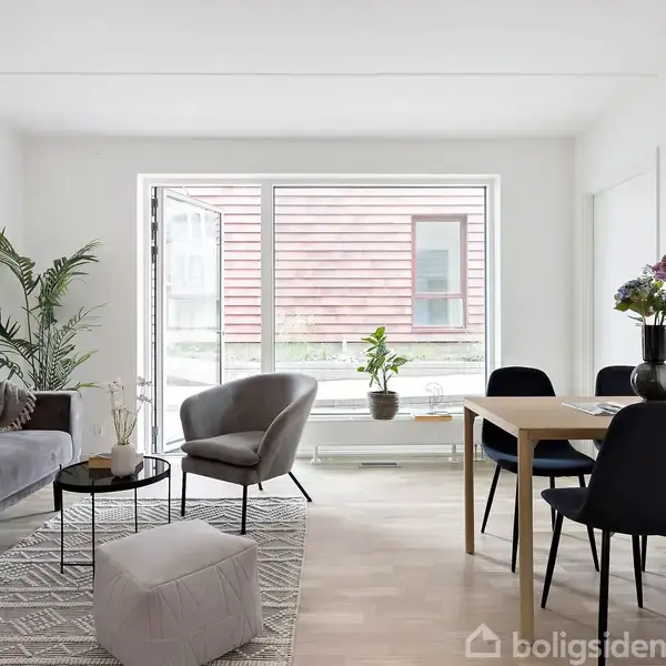 Stue med sofa og lænestol ved et sofabord. Spisebord med stole i nærheden. Store vinduer i baggrunden lader lys ind. Plante og blomsterdekorationer skaber en hyggelig atmosfære.